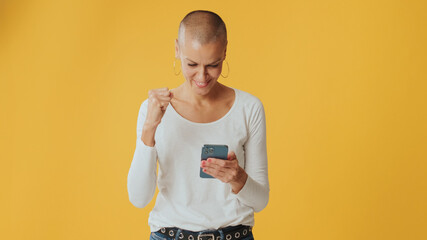 Smiling hairless woman, with mobile phone in hands celebrates victory, isolated on yellow...