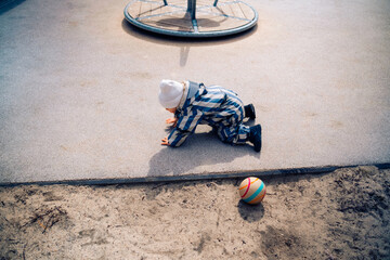 A boy child in striped overalls falls while playing with a ball. Kid aged about two years