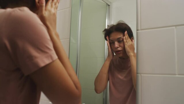 Zoom shot of young sleepy girl looking at her reflection in bathroom mirror and rubbing puffy face after waking up in morning at home