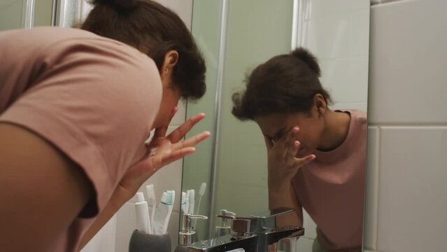 Drowsy teen girl washing her face with water above sink in bathroom and looking in mirror while feeling unwell and sleepy in morning at home