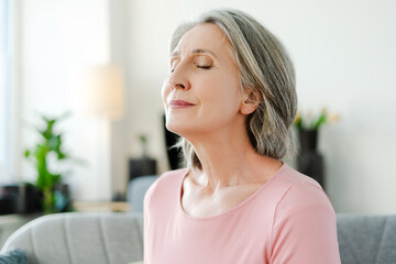 Smiling active senior woman sitting with closed eyes on comfortable sofa relaxing at home