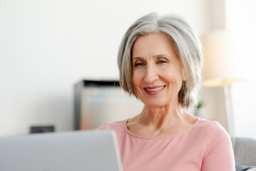 Attractive smiling senior woman using laptop computer, sitting on comfortable sofa