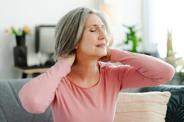 Unhappy senior woman stretching arms, doing exercises, having neck pain sitting on comfortable sofa