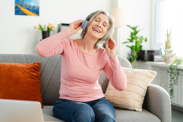 Happy senior woman listening music in wireless headphones, sitting on sofa