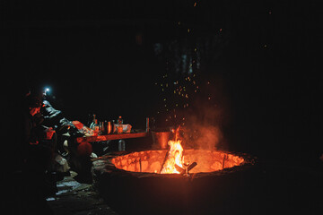 A diverse group of individuals stand in a circle around a crackling fire pit, sharing stories and...