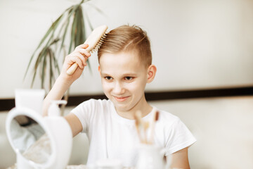 Smiling blonde teenager kid boy brushing hair with comb at home looking in small mirror. Morning...