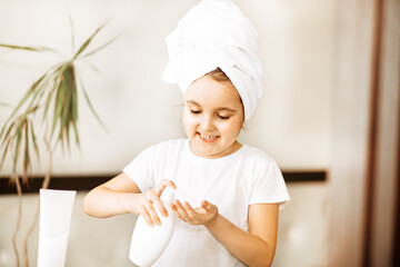 Funny little girl with towel on her head uses facial cream. Spa, healthcare, morning routine...