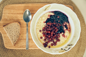 Top view of a plate of wheat porridge for a healthy gluten-free breakfast, next to a spoon and a piece of bread. A plate of wheat porridge with red currants and blueberry jam. High quality photo