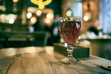 A crystal clear elegant glass of traditional multifaceted shape filled with wine stands on a wooden bar counter against a blurred background of the restaurant with a space to copy. High quality photo