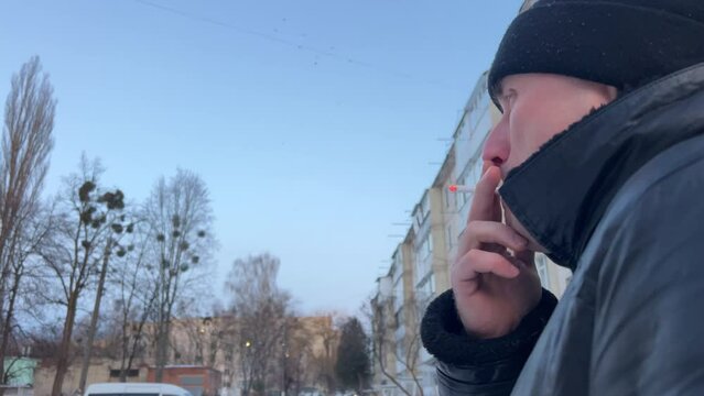 Close-up isolated portrait of man who smokes a cigarette and releases smoke into the camera lens. Bearded middle-aged man with bad habits smokes on the street. winter day.