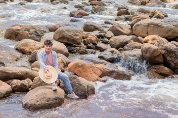 lifestyle. young latin guy sitting on the rocks by the river with hat in hand
