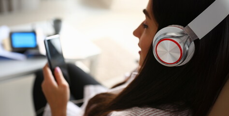 Young beautiful woman headphones holds smartphone in hand using at home costs about sofa. Verifies...