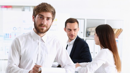 Handsome smiling bearded man in office look in camera with colleagues in background. White collar worker modern lifestyle graduate college study report idea coach train visit make note concept - obrazy, fototapety, plakaty