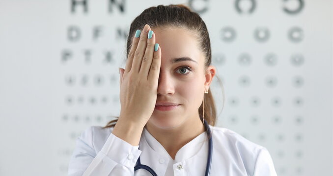 Portrait of gorgeous woman ophthalmologist standing in clinic office and covering right eye with tender hand. Lady looking at camera with happiness. Eyesight check concept