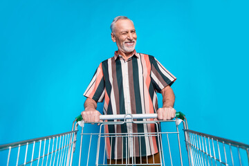 Photo of positive funky pensioner man wear summer striped trendy clothes hold cart isolated on blue...