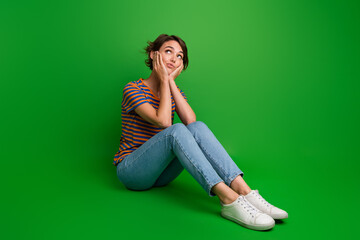 Full size portrait of creative nice girl sit floor arms touch cheeks look empty space contemplate isolated on green color background