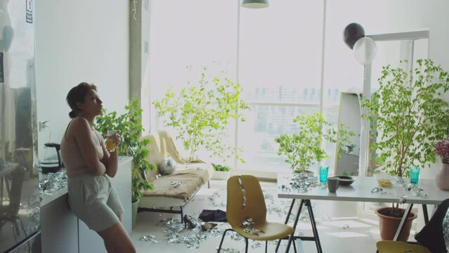 Young tired girl walking in messy living room with tinsel on floor, drinking juice and checking messages on mobile phone in morning at home