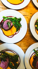 Many plates of beef tenderloin with sweet potato puree and green vegetables on a table awaiting servers to deliver the dishes. 