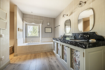 A spacious bathroom with wooden furniture with a double sink, black marble, two mirrors and a bathtub with vintage-style taps and white aluminum window and views of the forest