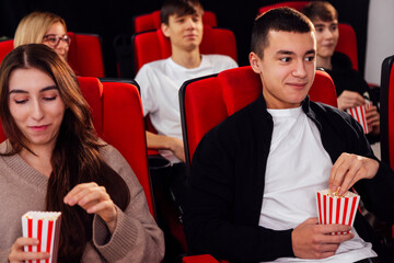 Nice young people in the cinema hall. Cute girls and boys are sitting in chairs and eating popcorn. Friends are watching a movie and laughing.