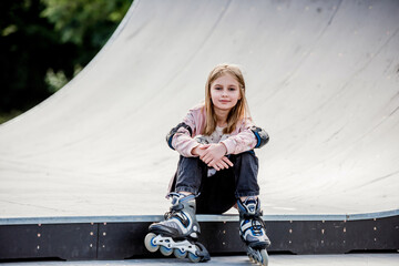 Cute girl roller skater sitting in city park and looking at camera Pretty female preteen kid child...