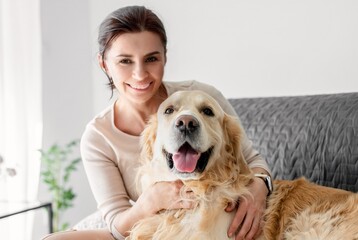 Girl hugging golden retriever dog at home and smiling looking at camera. Young woman with purebred pet doggy on sofa