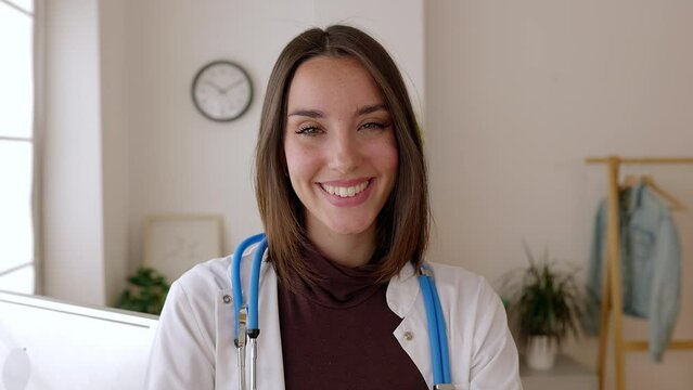 Smiling portrait of young female doctor standing at consultation room. Hospital worker in white coat looking at camera at clinical desk workplace. Healthcare people concept