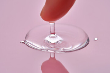 A finger touches a drop of cosmetic product on a pink background.