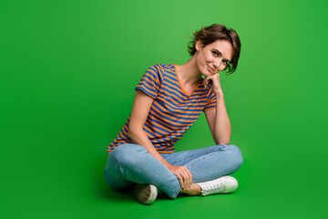 Full size portrait of lovely cheerful girl sit floor crossed legs hand touch chin isolated on green color background