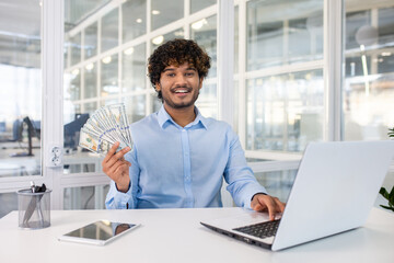 Successful financial analyst waving fan of dollar bills and looking at camera by desktop with...