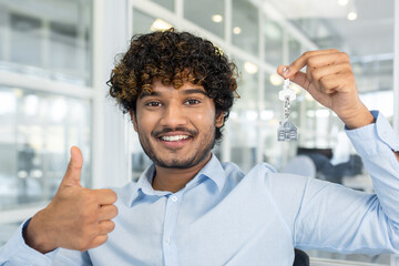 Diverse real estate agent smiling at camera while showing thumb up gesture and holding keys with...