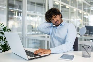 Struggling latino man rubbing neck and bending head while sitting by desktop with squinted face....