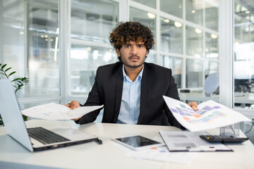 Frustrated indian male in jacket over shirt sitting by loaded table and spreading hands with documents. Young manager feeling stressed with paperwork and looking at camera with angry expression. - obrazy, fototapety, plakaty