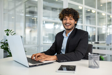 Arab businessman keeping hands on keyboard of portable computer while looking at camera with toothy smile. Curly employer feeling ready starting new productive and profitable week in office. - obrazy, fototapety, plakaty