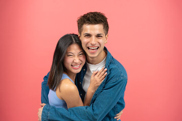 Fototapeta premium Loving young man hugging his pretty girlfriend on pink studio background, happy multiracial lovers enjoying time together, hugs, copy space.
