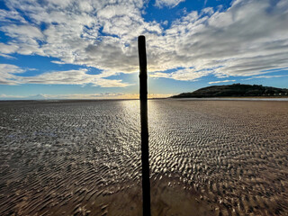 A wooden pole stands in the sand while the tide is out under a cloudy blue sky