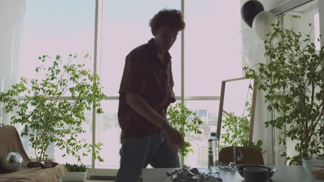 Young exhausted guy standing up fro couch, walking in living room, cleaning mess and drinking water while suffering from hangover after party at home