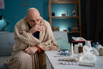 A man wrapped in a blanket suffering from chills and runny nose sitting on the sofa at home with medicine in the foreground