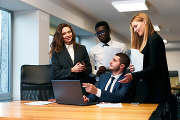 Multiethnic business team collaborates in modern office. African, Arabic, Caucasian professionals discuss strategy, celebrate success around laptop. Men, women in corporate attire engage in teamwork.