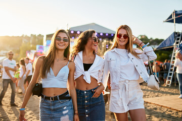 Three young women walk together on sunny beach at music festival. Happy friends in trendy summer...