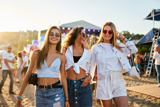 Three young women walk together on sunny beach at music festival. Happy friends in trendy summer outfits enjoy live concert, dancing, laughing in festive atmosphere. Group of girls having fun by sea.
