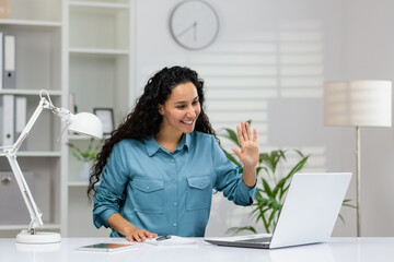 Distant dark haired recruiter in denim shirt waving hello to monitor of portable computer while sitting in light apartment interior. Friendly manager greeting candidates before beginning of interview.