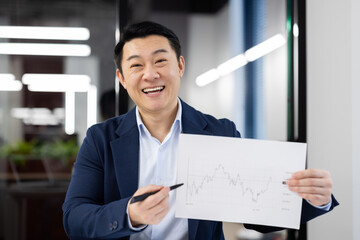 Cheerful taiwan investor showing sheet of paper with growing statistics and pointing with pen to...
