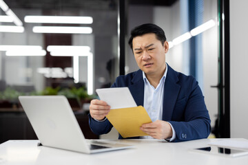 Disturbed asian man in business suit taking out letter from envelope while sitting in cabinet with...