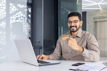 Fototapeta premium Positive unshaved male in eyewear looking at debit card while keeping hand on keyboard of modern computer. Carefree office worker enjoying possibility of making online payments in marketplaces.
