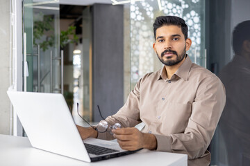 Fototapeta premium Serious islamic businessman taking off glasses during working day in personal workplace with computer. Concentrated startup runner making hard decisions about business growing strategy.