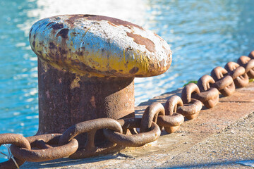 Cleat for mooring boats on concrete platform against a water background with rusty metal chain