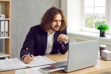 Portrait of a serious bearded young business man accountant in suit and long hair working at the...