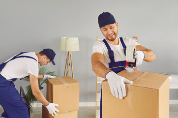 Moving and teamwork in transport services. Two professional young male movers securing cardboard boxes with adhesive tape in new home. Two male moving service workers in uniform carefully pack boxes.