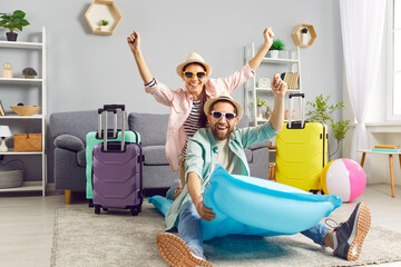 Playful funny couple having fun on air mattress at home, imagining themselves on summer vacation. Man and woman in summer hats and sunglasses are having fun at home among colorful suitcases.